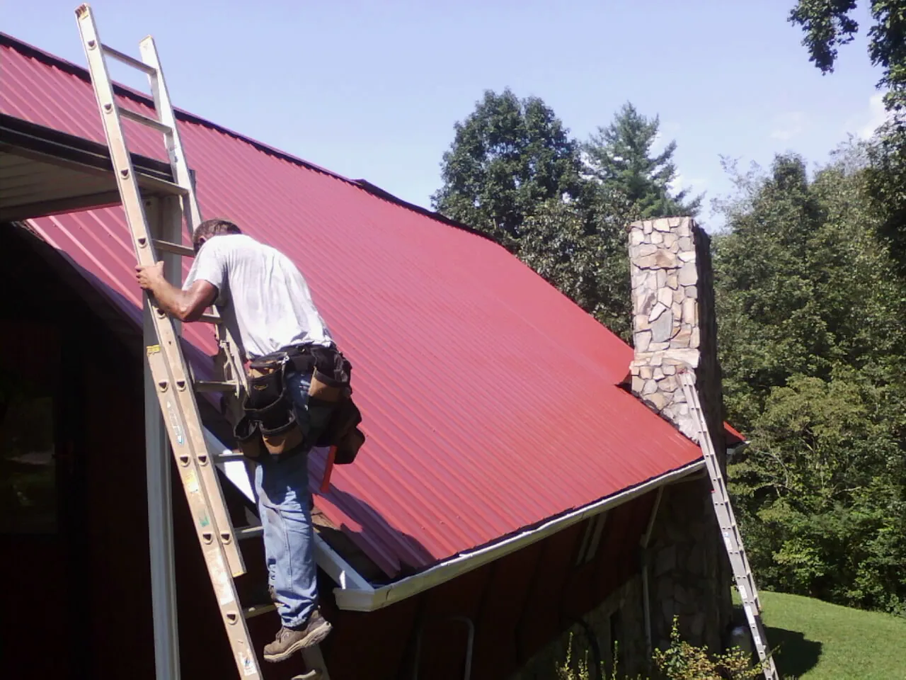Roofer on a ladder beside a red metal roof and stone chimney — Soffit & Fascia Repair by Roofing Brothers in Inchelium
